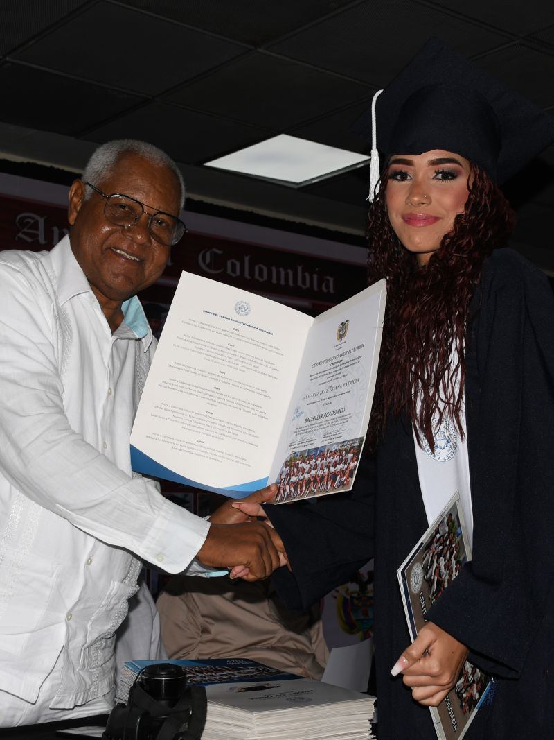 Graduada recibiendo su diploma de manos de un directivo, frente a un cuadro con fotos de promociones anteriores